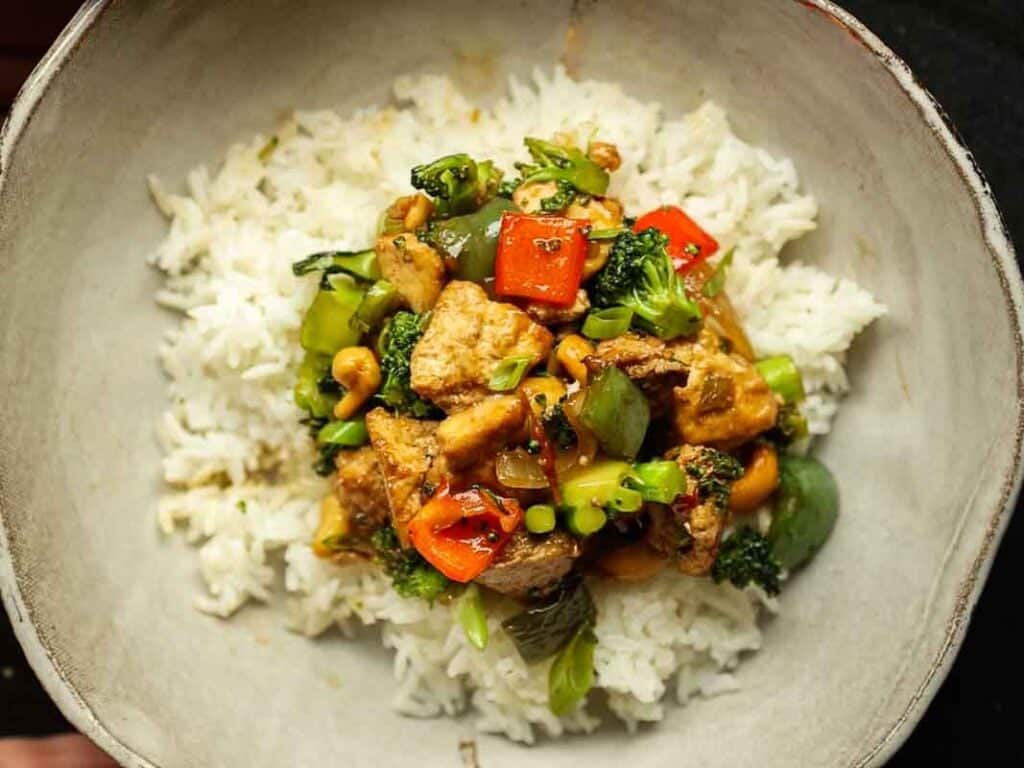 A bowl of white rice topped with a colorful cashew nut stir fry featuring pieces of tofu, red and green bell peppers, broccoli, and cashews.